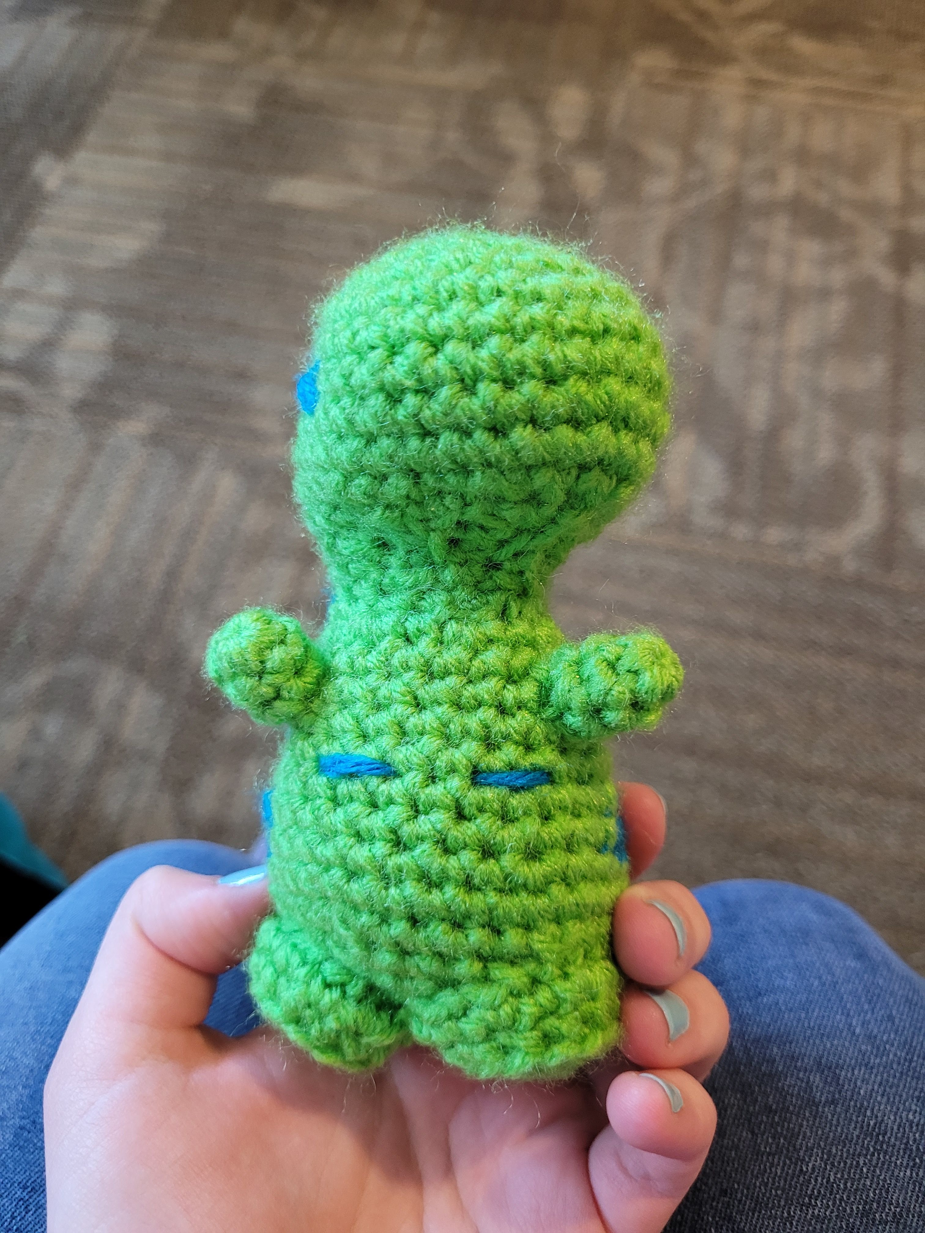 Green and blue crochet dinosaur from the front. It has top surgery scars sewn on.